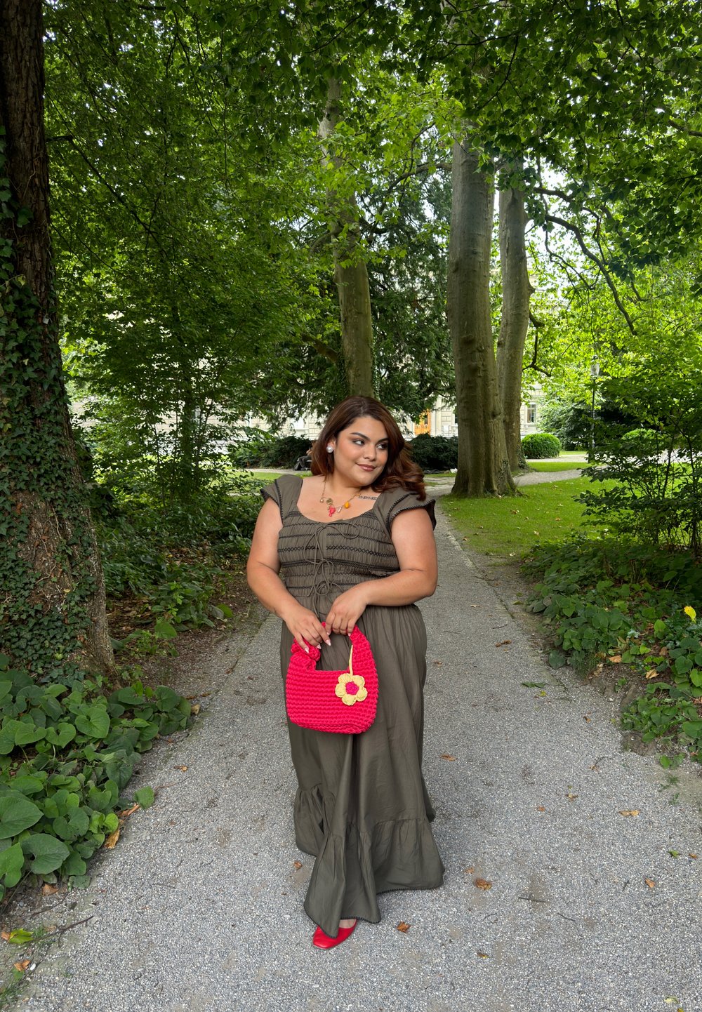 Ελιά πράσινο, tiered maxi φόρεμα με κοντά μανίκια και κορδόνι στη μέση; κρατώντας μια φωτεινή ροζ τσάντα χειρός με λουλούδι διακοσμητικό; κόκκιν α παπούτσια.