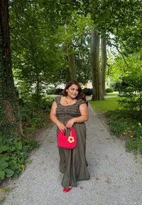 Ελιά πράσινο, tiered maxi φόρεμα με κοντά μανίκια και κορδόνι στη μέση; κρατώντας μια φωτεινή ροζ τσάντα χειρός με λουλούδι διακοσμητικό; κόκκινα παπούτσια.