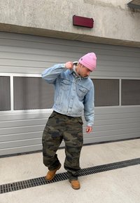 Jasnoniebieska kurtka dżinsowa, szary hoodie pod spodem, spodenki cargo w kamuflażu, beżowe buty oraz różowa czapka. Stoi w tle szarego garażu.