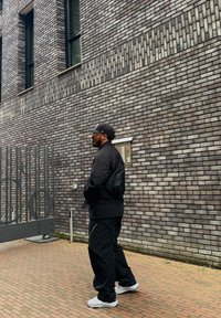 Black bomber jacket, black trousers, and white trainers. A grey brick wall in the background, featuring large windows and a metal gate.