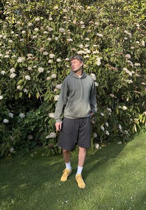 Joven con sudadera gris con capucha, pantalones cortos negros y zapatos amarillos, de pie sobre el césped frente a un arbusto frondoso con flores blancas.