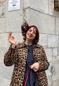 Leopardenmuster Mantel mit Knopfverschluss, getragen über einem dunklen Kleid. Die Frau hält mit einer Hand einen roten Riemen und steht vor einer Steinmauer.