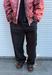 Rdeča bomber jakna z zadrgo spredaj, črne hlače širokih hlačnic s črtami in rjavi čevlji z brušenim zaključkom, stoji pred svetlo ozadje.