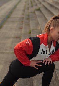 Ženska v rdeči, črni in beli Nike jakni razteguje nogo na zunanjih betonskih stopnicah in se pripravlja na vadbo.