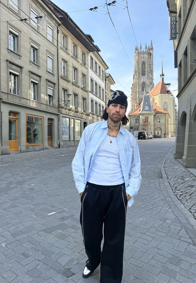 Muž s tetováním, oblečený v bílém tričku, černých kalhotách a kulichu, stojí na dlážděné ulici s historickými budovami a kostelní věží za ním.
