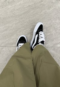 Μαύρα και λευκά αθλητικά παπούτσια με λευκές γλώσσαν, συνδυασμένα με ένα μακρύ, ελιόχρωμο ύφασμα. Το πάτωμα είναι ανοιχτό γκρίζο, με λεία υφή.