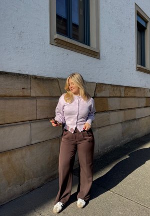 Mujer rubia con cárdigan lavanda y pantalones marrones posa contra una pared de piedra, sosteniendo gafas de sol, con la luz del sol proyectando una sombra en el pavimento.