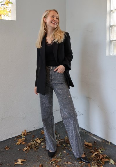 Blazer negro sobre una blusa negra, combinado con pantalones de pierna ancha de talle alto con lentejuelas plateadas y tacones negros, con una pared clara de fondo.