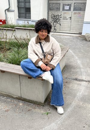 Chaqueta beige clara con cuello marrón, jeans azul oversized y zapatillas blancas. Bolso cruzado negro. Cabello rizado, sentado en un banco de madera.