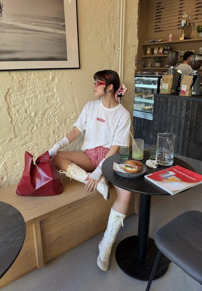 Bolso de cuero rojo, camiseta blanca de gran tamaño con el texto "¿CÓMO?", pantalones cortos de cuadros rojos, botas altas blancas con cordones y gafas de sol. Mesa con una hamburguesa.