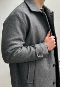 Manteau en laine grise avec col structuré, poignets boutonnés et coupe élégante et ajustée. Présente une texture subtile et des boutons foncés.