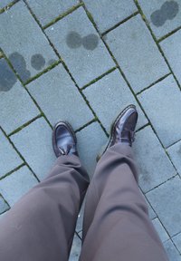 Tumed pruun nahast loafers fringi detailiga, kollased õmblused ja läbipaistvad tallad, paigutatud hallidele sillutuskividele, millel on sammal.