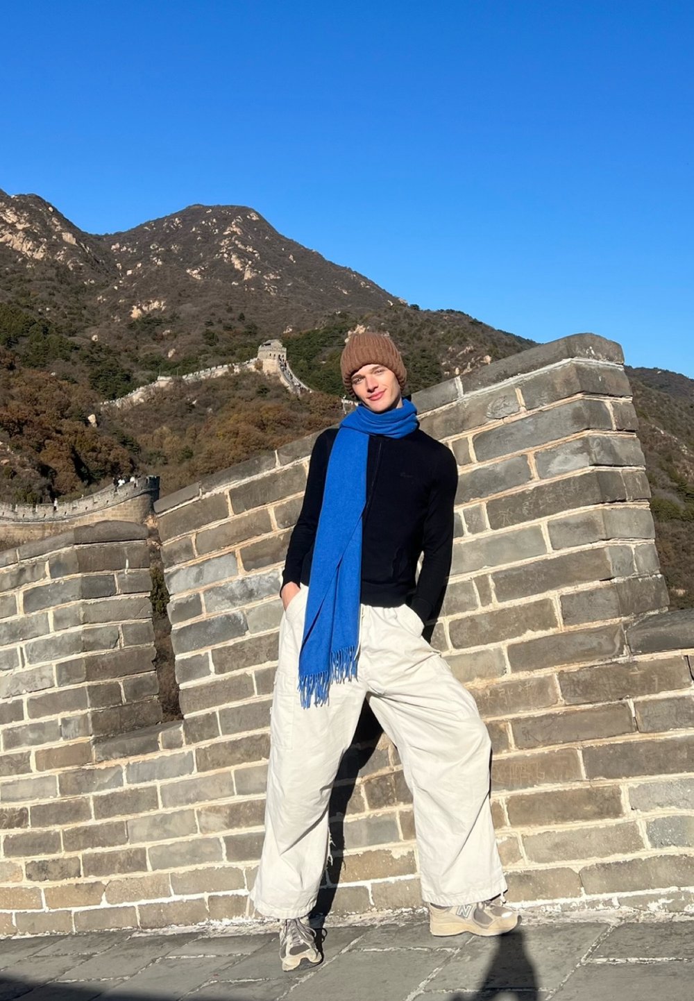 Oseba, ki nosi črno majico z dolgimi rokavi, bež široke hlače, bež superge in rjavo kapa, z modrim šalom, stoji ob kamnitem zidu.