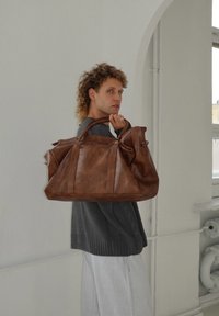 Mala de viagem de couro castanho com forma estruturada, costuras proeminentes e alças duplas. Usada casualmente ao ombro, contra um fundo claro.