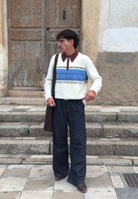 Biały puszysty sweter w niebieskie i brązowe paski, brązowy kołnierz; luźne ciemne dżinsy; brązowy pasek; czarna torba na ramię; stojący na kamiennych schodach.