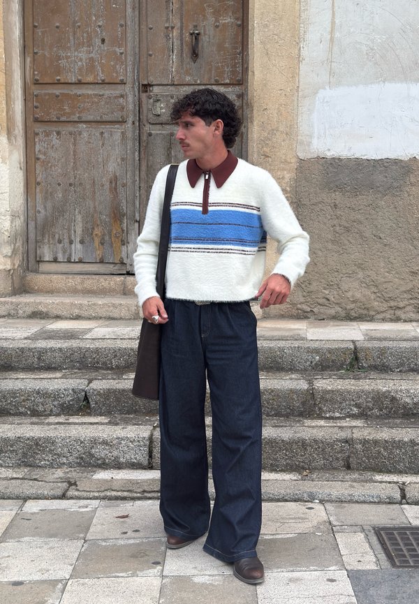 Weißer, flauschiger Pullover mit blauen und braunen Streifen, braunem Kragen; lockere, dunkle Jeanshose; brauer Gürtel; schwarze Schultertasche; steht auf Steinstufen.