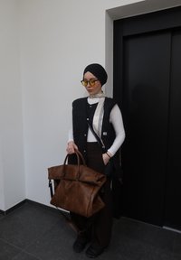 Femme portant un turban noir et un gilet, lunettes teintées jaunes, tenant un grand sac en cuir marron, debout près de portes d'ascenseur noires fermées.