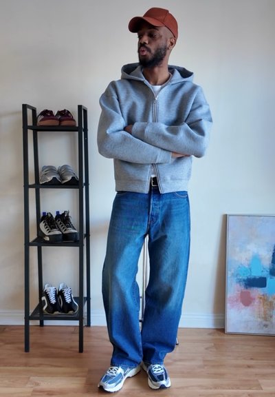 Hombre con vaqueros azules, sudadera gris con capucha y gorra marrón, de pie con los brazos cruzados cerca de un zapatero y una pintura abstracta en un suelo de madera.