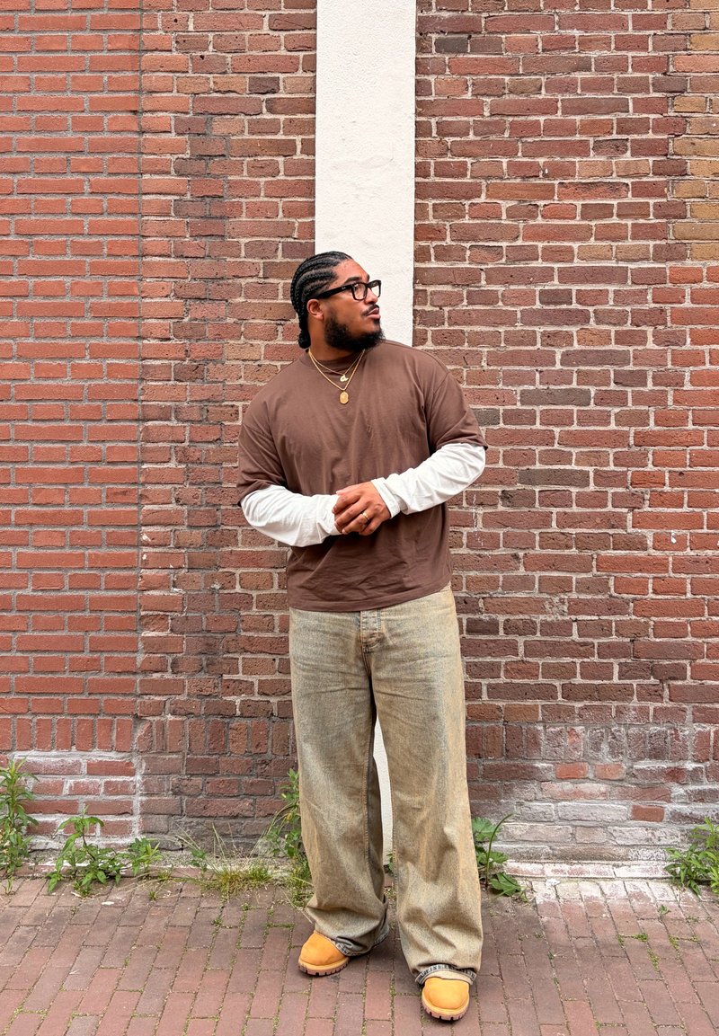 Homme aux cheveux tressés portant des lunettes, une chemise marron sur des manches longues blanches, un pantalon beige et des chaussures jaunes, debout contre un mur de briques avec de petites plantes.