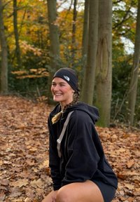 Svart hettegenser, svart pannebånd og shorts; sitter på et løvdekket underlag i et skogsområde med høstløv i bakgrunnen.