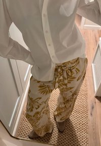 Camisa blanca abotonada combinada con pantalones sueltos color crema con un estampado floral amarillo y cintura con cordón, sobre una alfombra tejida.