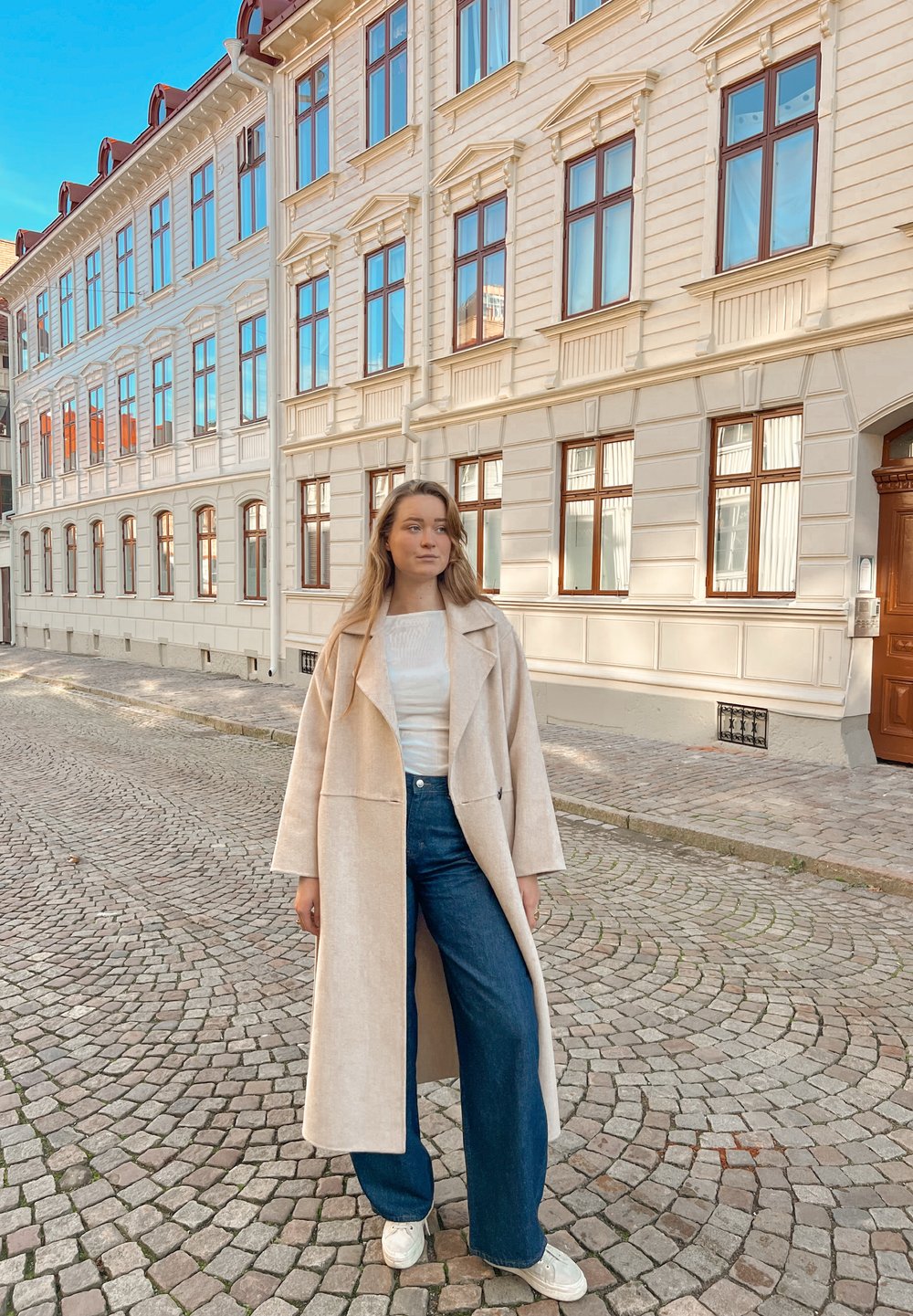 Long manteau beige sur un haut blanc, associé à un jean bleu à jambes larges et des baskets blanches. Rue pavée et bâtiments historiques en arrière-plan.