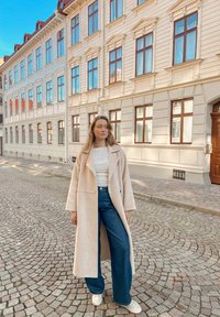 Long manteau beige sur un haut blanc, associé à un jean bleu à jambes larges et des baskets blanches. Rue pavée et bâtiments historiques en arrière-plan.