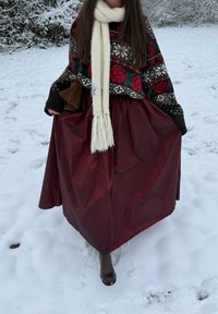 Kvinde i sne iført mønstret sweater, cremefarvet tørklæde, lang bordeaux nederdel, brune støvler, holder nederdelens kanter og en brun håndtaske.