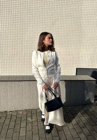 Biała dziana kardigan z guzikami, nałożony na kremową sukienkę. Czarna torebka oraz czarne buty z białymi skarpetkami dopełniają stylizację. Tło w kwadratowe płytki.