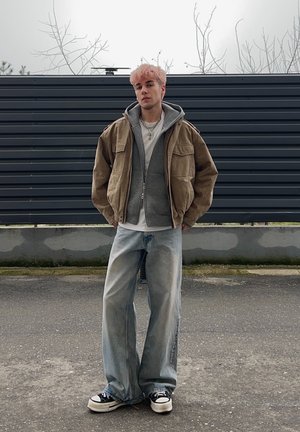 Persona joven con cabello rosa que lleva una chaqueta marrón sobre una sudadera gris, camiseta blanca, jeans anchos y zapatillas negras y blancas, de pie al aire libre.