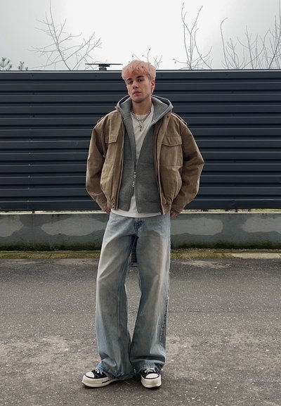 Persona joven con cabello rosa que lleva una chaqueta marrón sobre una sudadera gris, camiseta blanca, jeans anchos y zapatillas negras y blancas, de pie al aire libre.