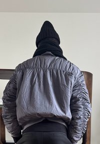 Personne vue de dos portant une veste grise matelassée, un bonnet en tricot noir et une écharpe noire à l'intérieur, avec des meubles en bois en arrière-plan.