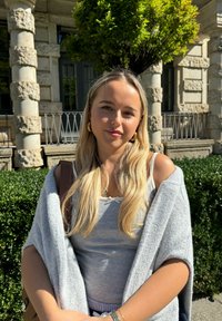 Szary kardigan na jasnoszarym topie na ramiączkach, luźny krój, z długimi falującymi blond włosami. W tle zielone liście i kamienny budynek.