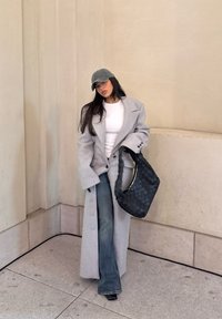 Grey oversized coat, white top, blue flared jeans, and grey cap. Black woven handbag hangs from shoulder. Neutral background.
