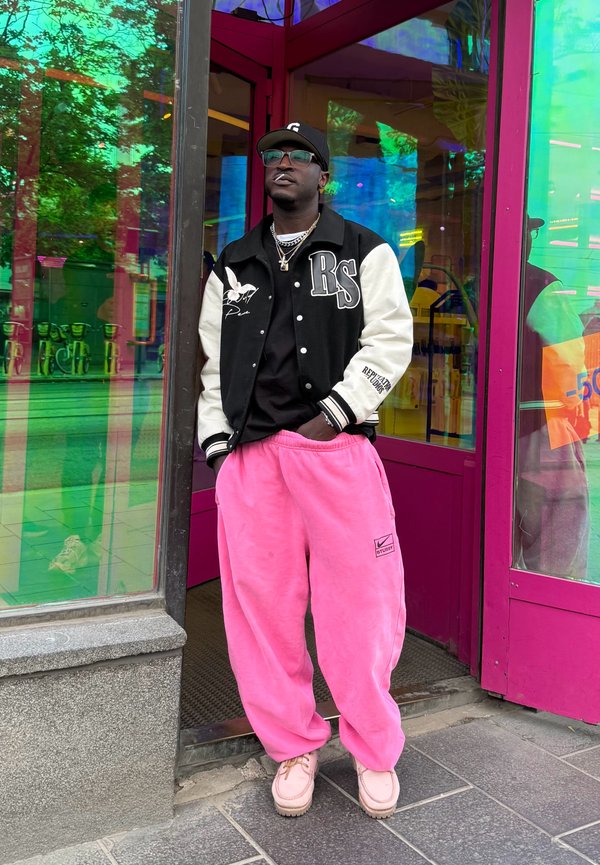 Mann, der eine schwarz-weiße Collegejacke, pinke Hosen, beige Schuhe und eine schwarze Kappe trägt, steht vor einer pinken Glastür mit Spiegelungen.