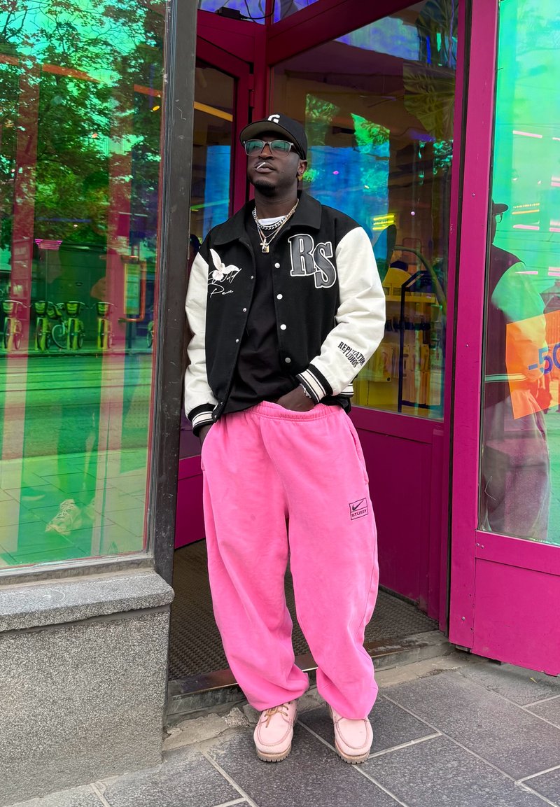 Homme portant une veste varsity noire et blanche, un pantalon rose, des chaussures beiges et une casquette noire, debout devant une porte en verre rose avec des reflets.