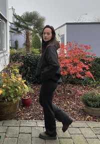 Juodas, perdėtas švarkas su aukštu apykakle, derinamas su juodomis kelnėmis ir tamsiais batais. Lauko aplinka su augalais ir rudens lapais.