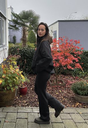 Chaqueta negra de gran tamaño con un alto cuello, combinada con pantalones negros y botas oscuras. Entorno al aire libre con plantas y follaje otoñal.