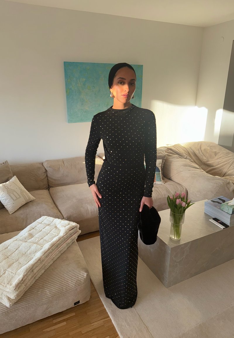 Femme en longue robe noire à petits pois, foulard noir, tenant un chapeau noir, debout dans un salon beige avec des fleurs sur une table.