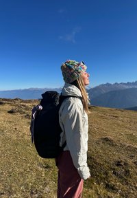 En svart ryggsekk med klaff på toppen, båret på ryggen, kombinert med en lys grå jakke og en fargerik strikket lue. Fjellandskap i bakgrunnen.