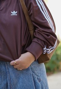 Brun Adidas sweatshirt med hvide striber, parret med en lyseblå denimnederdel. En brun skuldertaske hviler på skulderen, fingre griber fat i trøjen.