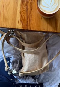 Tan fabric handbag with a textured surface, adjustable straps, and silver hardware resting on striped fabric, accompanied by a latte in a ceramic cup.