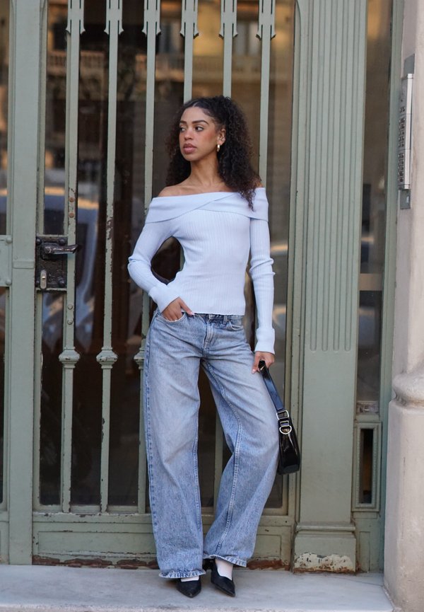 Svetlo modra rebrasta majica z odprtimi rameni, ohlapne sive kavbojke, črna torbica in črni koničasti čevlji pred zelenimi starinskimi vrati.