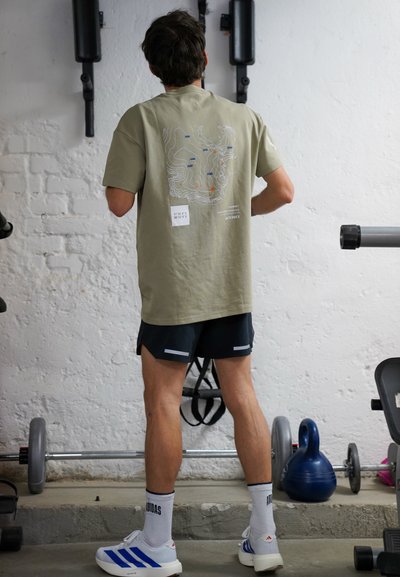 Hombre con camiseta caqui con diseño de mapa, pantalones cortos negros y zapatos blancos Adidas, de pie en un gimnasio cerca de pesas y pesas rusas.