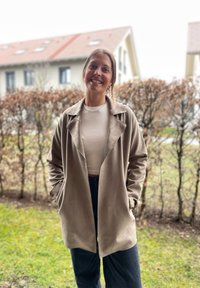 Bézs oversize dzseki sálgallérral és nagyméretű elülső zsebekkel, puha anyagból készült. Fehér ing és sötét nadr�ág fölé viselve.