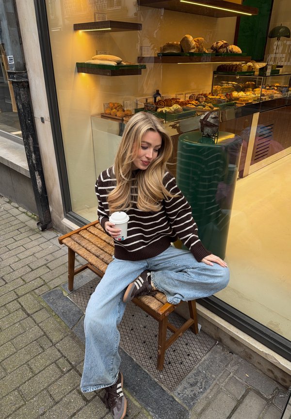 Ung kvinne i stripete genser og jeans sitter på benk utenfor bakeri og holder en kaffekopp, med ulike brød vist inne.