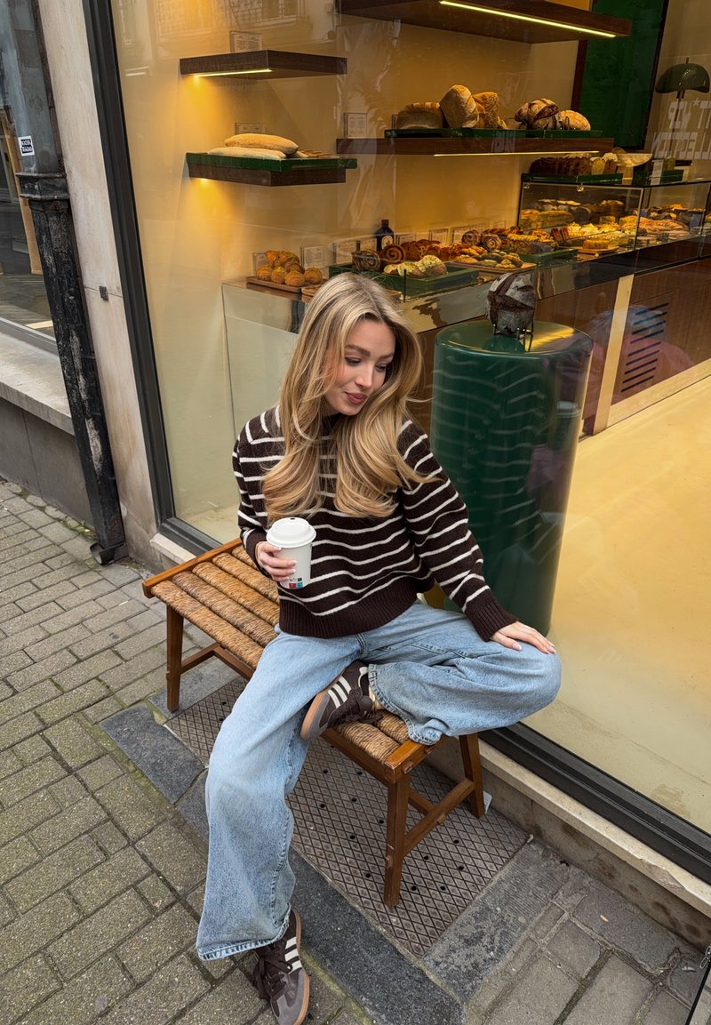 Ung kvinna i randig tröja och jeans sitter på bänk utanför bageri och håller en kaffekopp, med olika sorters bröd utställda inuti.