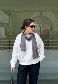 Hvid, fluffy sweater med ribbede manchetter, grå tørklæde lagt rundt om halsen, sorte solbriller, mørke bukser, stående mod en glasbaggrund.
