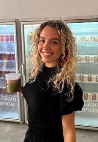 Svart teksturert kjole med puffermer; holder en klar kopp med lagdelt grønn og rosa drikk. Kjøleskap fylt med fargerike drikker i bakgrunnen.