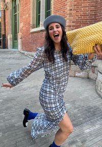 Szürke kockás ruha ráncolt dizájnnal, puffos ujjakkal és hasított szegéllyel. Világosszürke kalapot visel és egy szőtt sárga táskát tart a kezében.